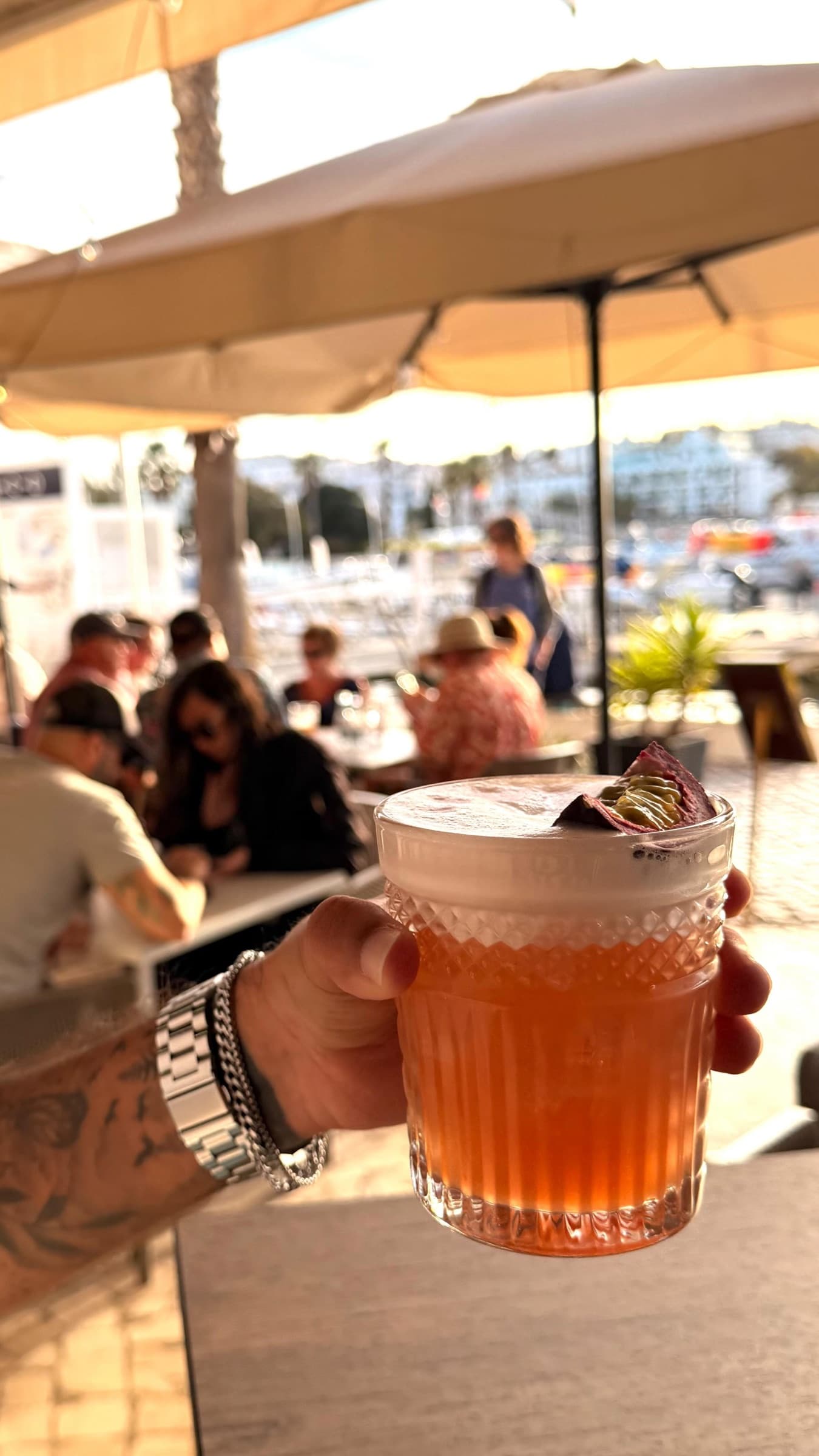 A cocktail held up on the busy terrace in the late sun, umbrellas and the marina behind